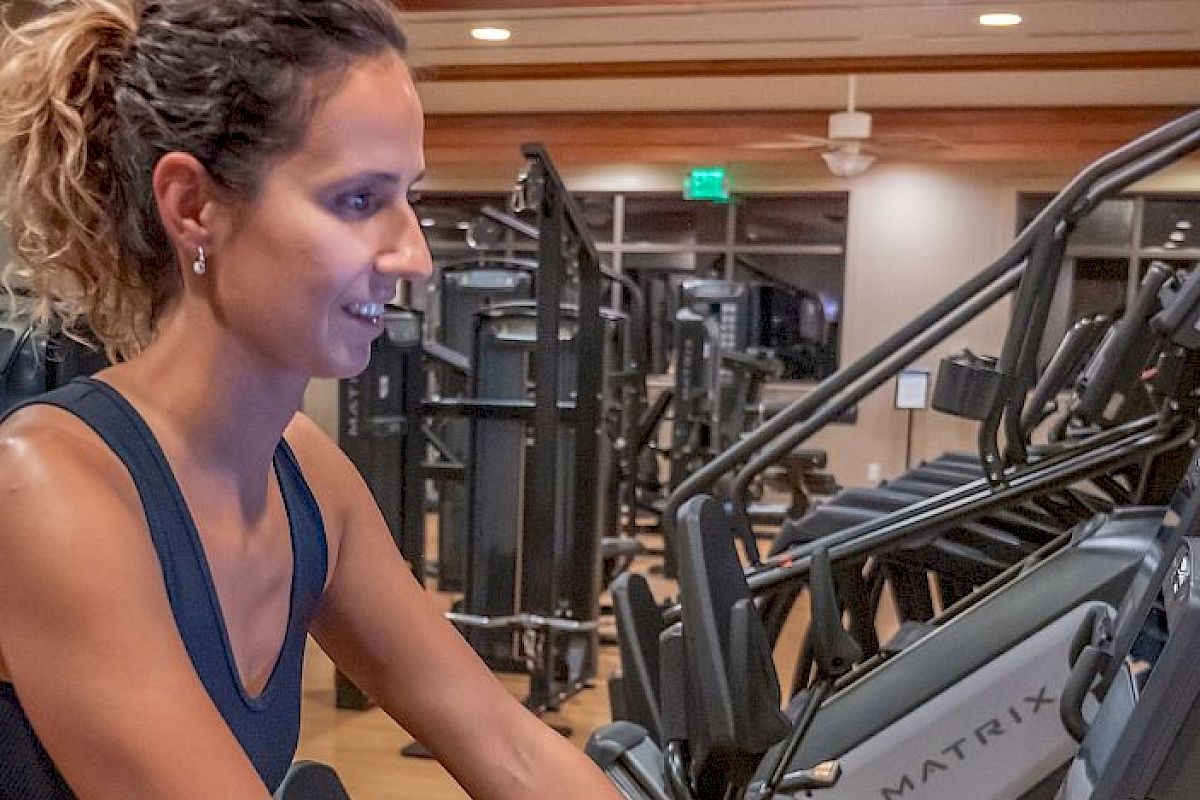 A woman exercises on a stationary bike in a gym, smiling and focused. She has a tattoo on her arm and is surrounded by various workout equipment.