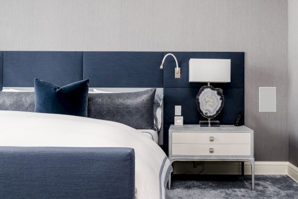 A modern bedroom with a blue and white color scheme, featuring a bed, pillows, a nightstand, a lamp, and a decorative item.