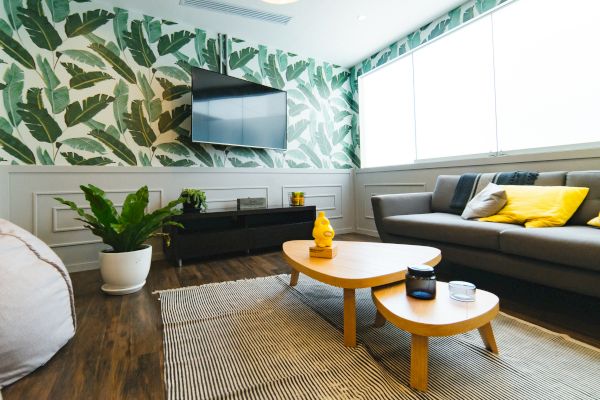 A modern living room with a tropical leaf wallpaper, a wall-mounted TV, a grey sofa with yellow cushions, and a wooden coffee table.