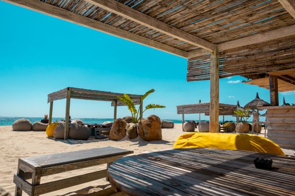 A serene beach scene with wooden shaded structures, comfy seating areas, and a view of the ocean under a clear blue sky.