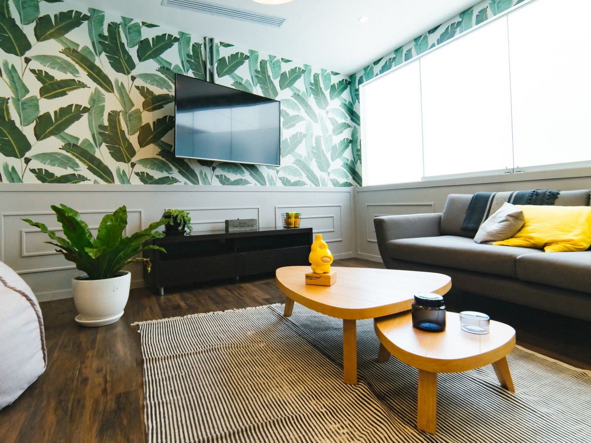 A living room with a tropical leaf wallpaper, a gray sofa with yellow cushions, a wooden coffee table, potted plants, and a wall-mounted TV.