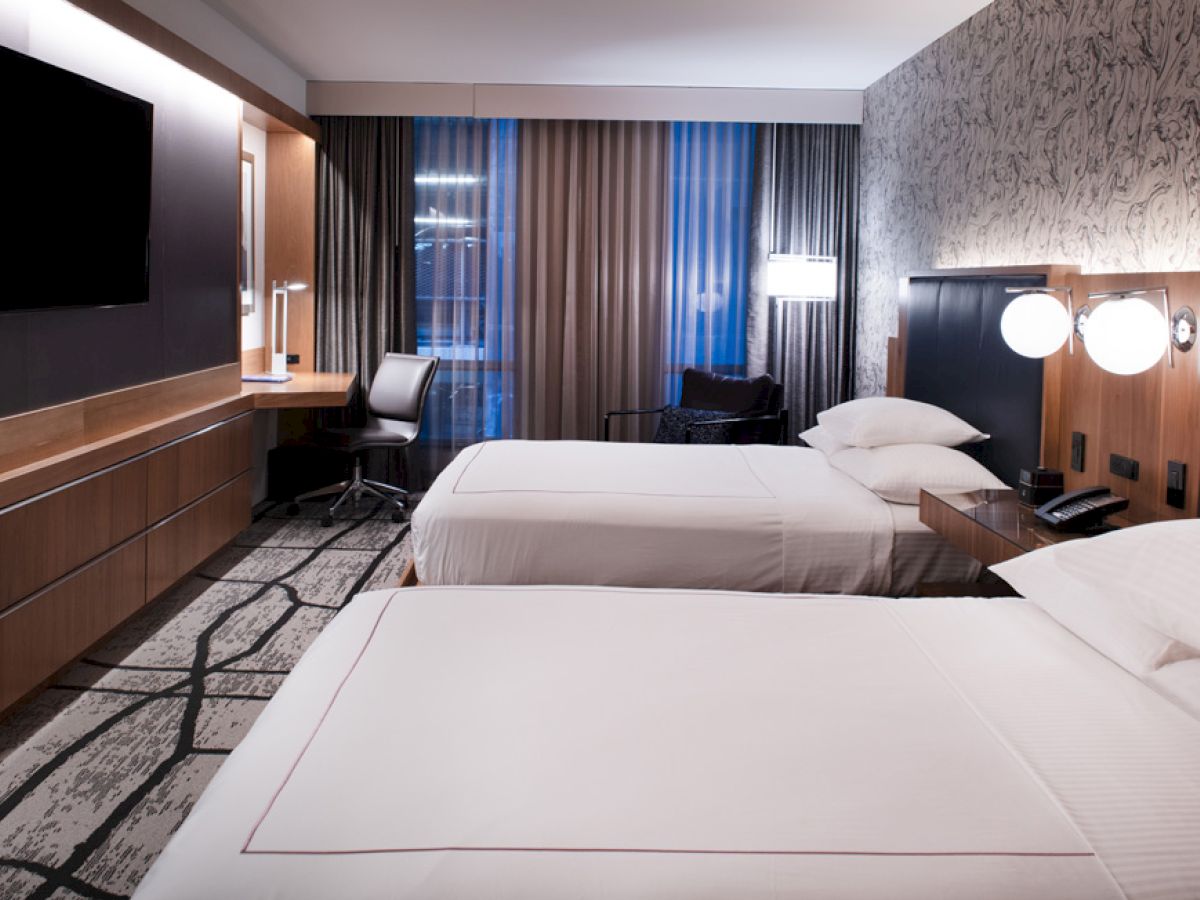 A modern hotel room with two neatly made beds, a desk, a large TV, and stylish decor featuring patterned walls and curtains, with ambient lighting.