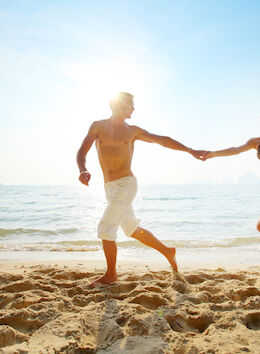 A couple is running hand-in-hand along a sandy beach near the ocean, with the bright sun shining behind them.