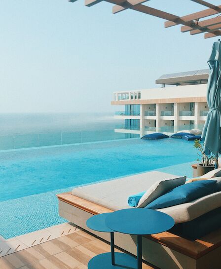 The image shows a luxury poolside area with lounge chairs, an umbrella, and a clear view of the ocean in the background.