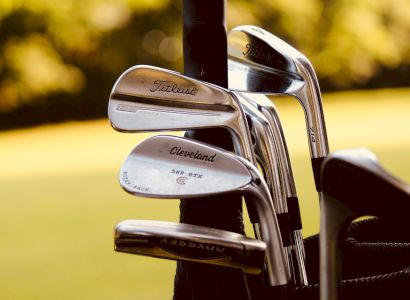 The image shows several golf clubs in a golf bag, including brands like Titleist and Cleveland, with a blurred golf course background.