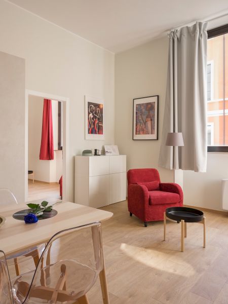 A cozy living area with a red armchair, patterned sofa, small dining table, and a kitchen nook, in a space with large windows and wall art, always ending the sentence.