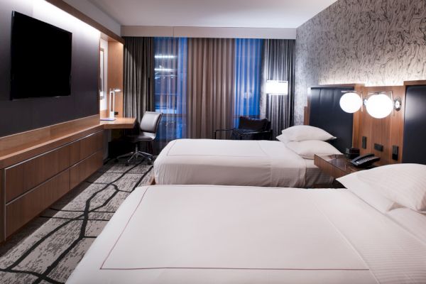 The image shows a modern hotel room with two beds, a desk with a chair, a flat-screen TV, and elegant lighting.