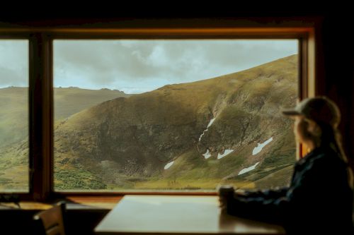 A person is sitting at a table by a window, gazing at a scenic mountain landscape with patches of snow on the slopes, bathed in natural light.
