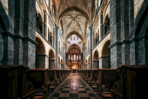 The image shows the interior of a grand cathedral with towering arches, rows of wooden pews, stone columns, and an illuminated altar at the far end.