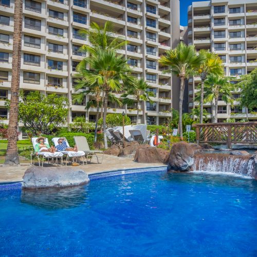 A swimming pool with a small waterfall feature, surrounded by lounge chairs, palm trees, and a multi-story building in the background.