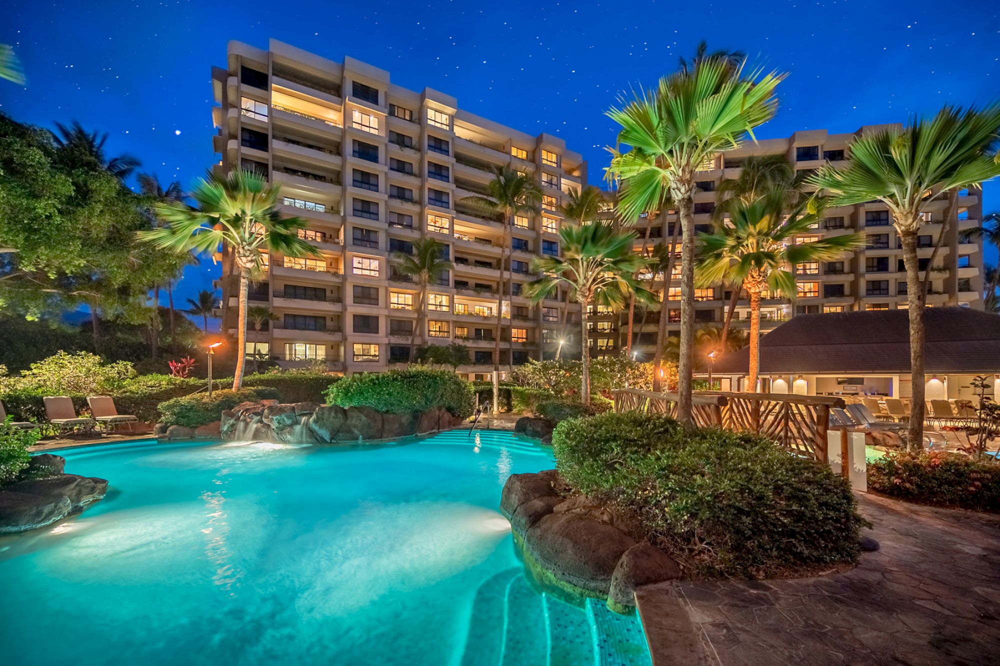 A luxurious resort with a lit-up swimming pool, surrounded by lush greenery and tall buildings, under a starry night sky.
