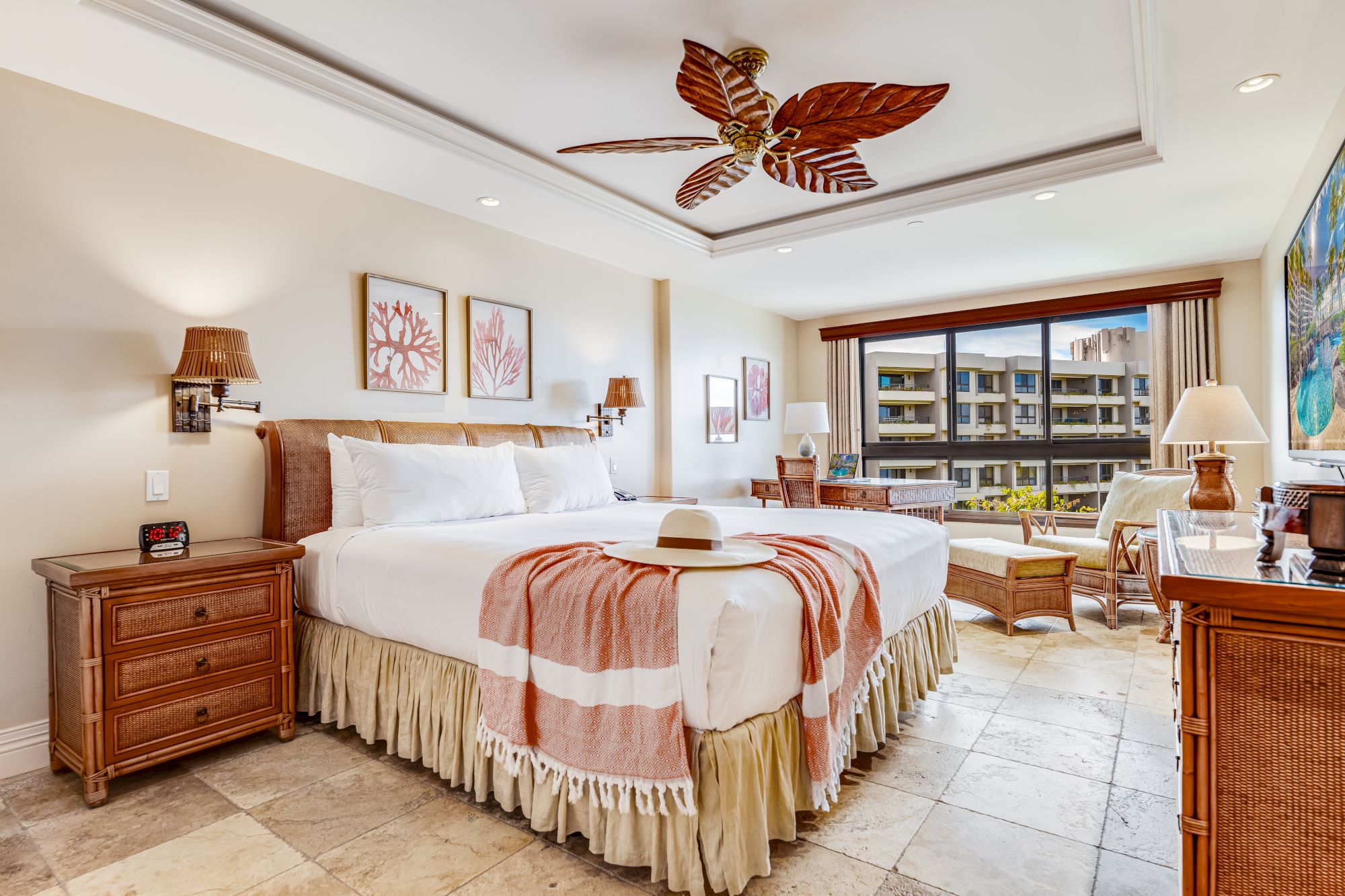 A hotel room with a large bed, two nightstands, lamps, and a ceiling fan. It has a balcony with a view of buildings and plants outside.