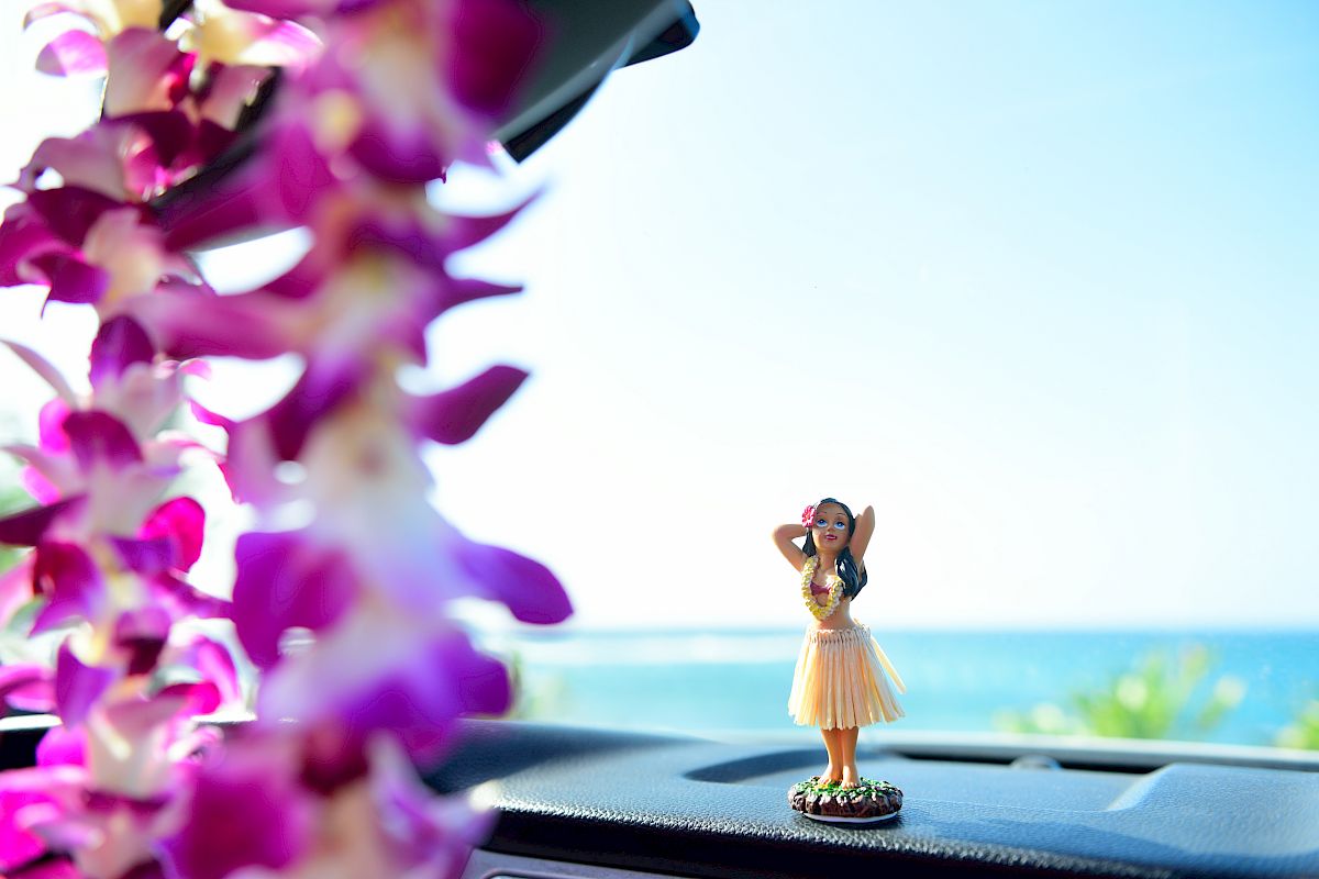 A hula dancer figurine sits on a car dashboard with a purple flower lei hanging, and the backdrop is a bright, sunny ocean view.