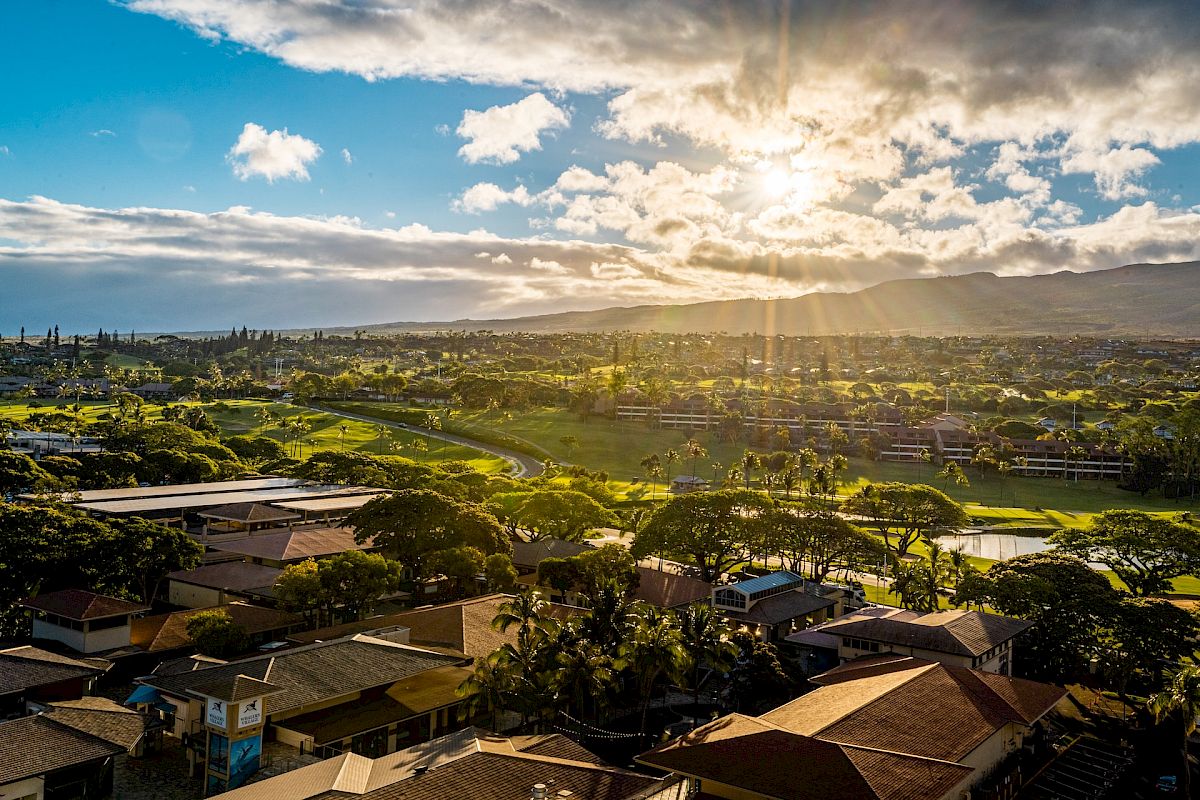 A picturesque aerial view of a town with houses nestled among green landscapes, set against a backdrop of mountains and a partly cloudy sky.