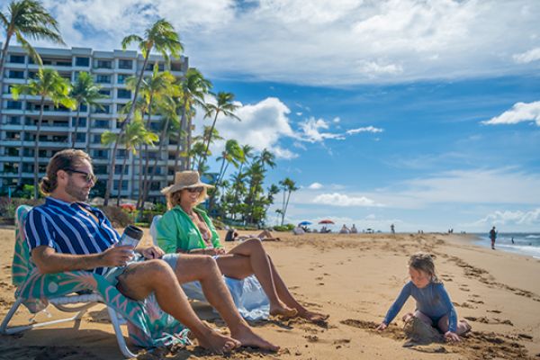 A couple relaxes in beach chairs under palm trees while a child plays in the sand, with a hotel and a clear blue sky in the background.