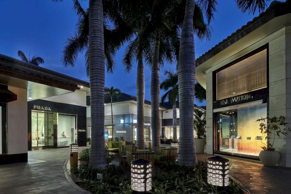 An outdoor shopping area with stores including Prada and Louis Vuitton, surrounded by palm trees and lit street lamps at dusk, providing a luxurious ambiance.
