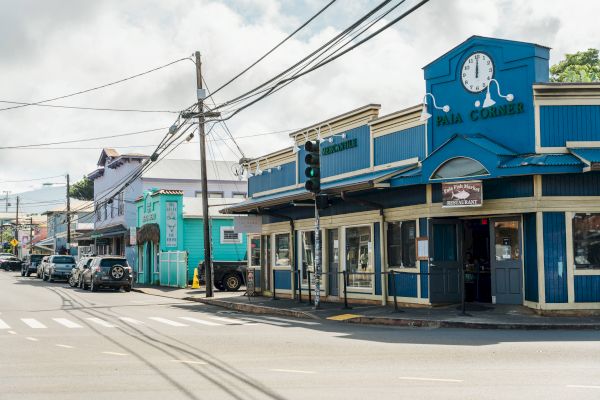 A street corner with colorful buildings, including a blue one named "Pāʻia Corner" with a clock on top, and cars parked along the road.