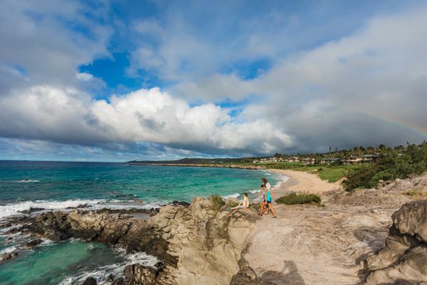 A couple walks along a rocky coastline with turquoise waters and a cloudy sky, possibly a rainbow forming in the distance, on a serene beach.