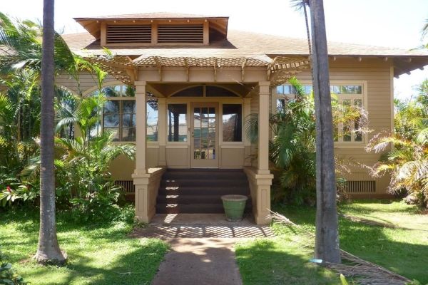 The image shows a yellow house with a covered porch and stairs leading up, surrounded by palm trees and greenery on a sunny day.