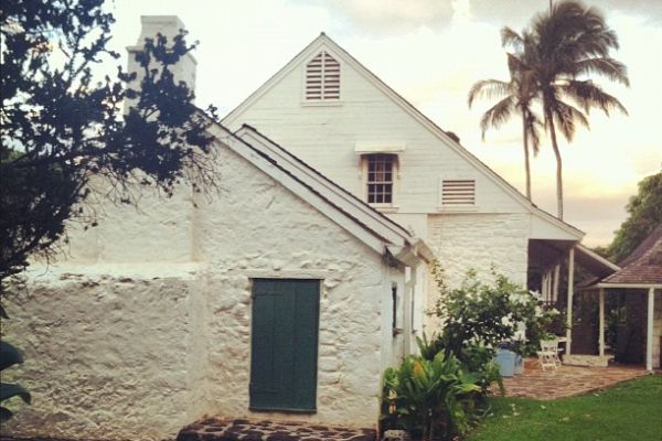 A white, rustic house with a green door is surrounded by stone pathways, lush greenery, and palm trees under a partly cloudy sky at dusk.