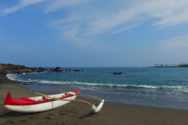 A small outrigger canoe with red and white colors is on a sandy beach with a calm ocean and partly cloudy sky in the background.