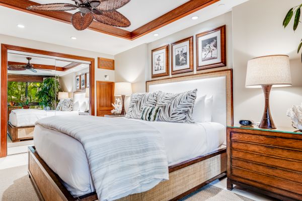 A well-decorated bedroom with a bed, tropical-themed pillows, lamps on nightstands, framed photos above the headboard, and a mirror reflecting the room.