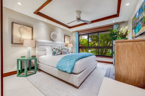 A modern bedroom with a large bed, turquoise accents, ceiling fan, window with lush greenery outside, and decorated with artwork and plants.