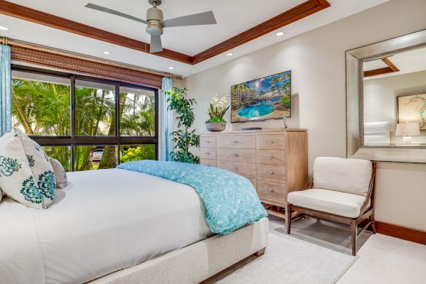 A cozy bedroom with a large bed, dresser, TV, chair, mirror, and window view of greenery. Room has ceiling fan and decorative plants, ending the sentence.