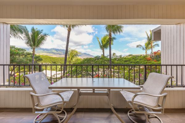 A balcony with two cushioned chairs and a table overlooks a lush landscape with palm trees and distant mountains under a blue sky.