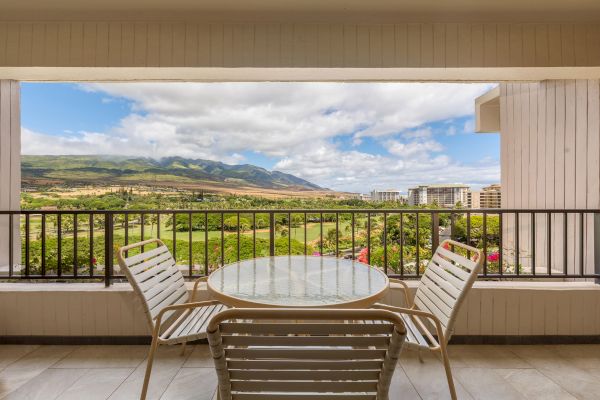 A balcony features a round glass table with four chairs, overlooking a scenic view of green fields, mountains, and distant buildings under a partly cloudy sky.