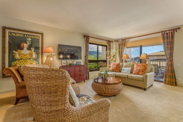 A cozy living room with wicker chairs, a round coffee table, a sofa, a TV, large windows with drapes, and a decorative wall poster.