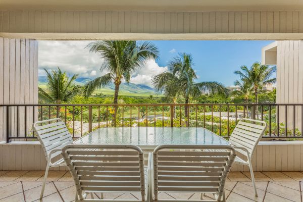 An outdoor patio area with a glass table and four white chairs overlooks a lush landscape with palm trees and mountains in the background.