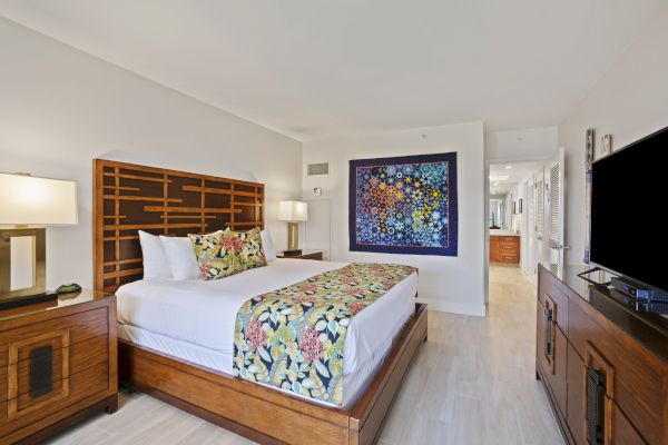 A hotel room with a king-sized bed, colorful artwork above, wooden furniture, two lamps, a TV on a dresser, and a hallway leading to another room.