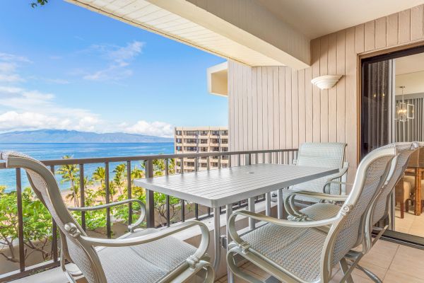 A balcony with a table and chairs overlooks a beachfront view with palm trees, a building, and an ocean horizon under a partly cloudy sky.