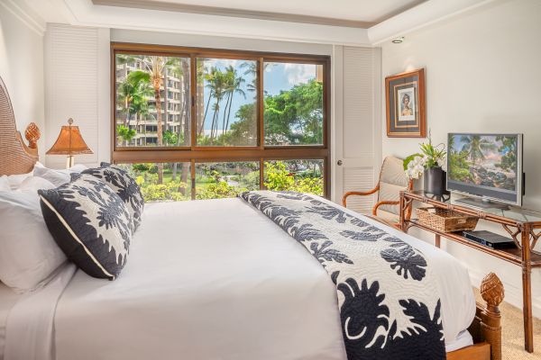 A bright room with a large bed, decorative pillows, a TV, a chair, and a view of lush greenery and buildings outside the large window.