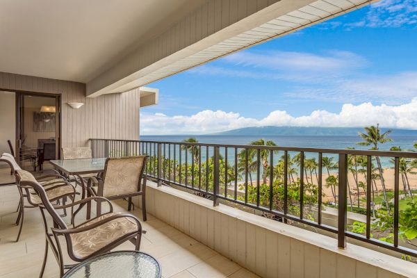 The image shows a balcony with chairs and a table, overlooking a beautiful beach and ocean with blue skies and palm trees in the background.