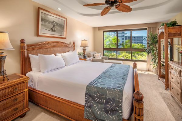 A spacious bedroom features a large wooden bed with white linens, bedside tables, lamps, ceiling fan, a couch, dresser, flat-screen TV, and large window.