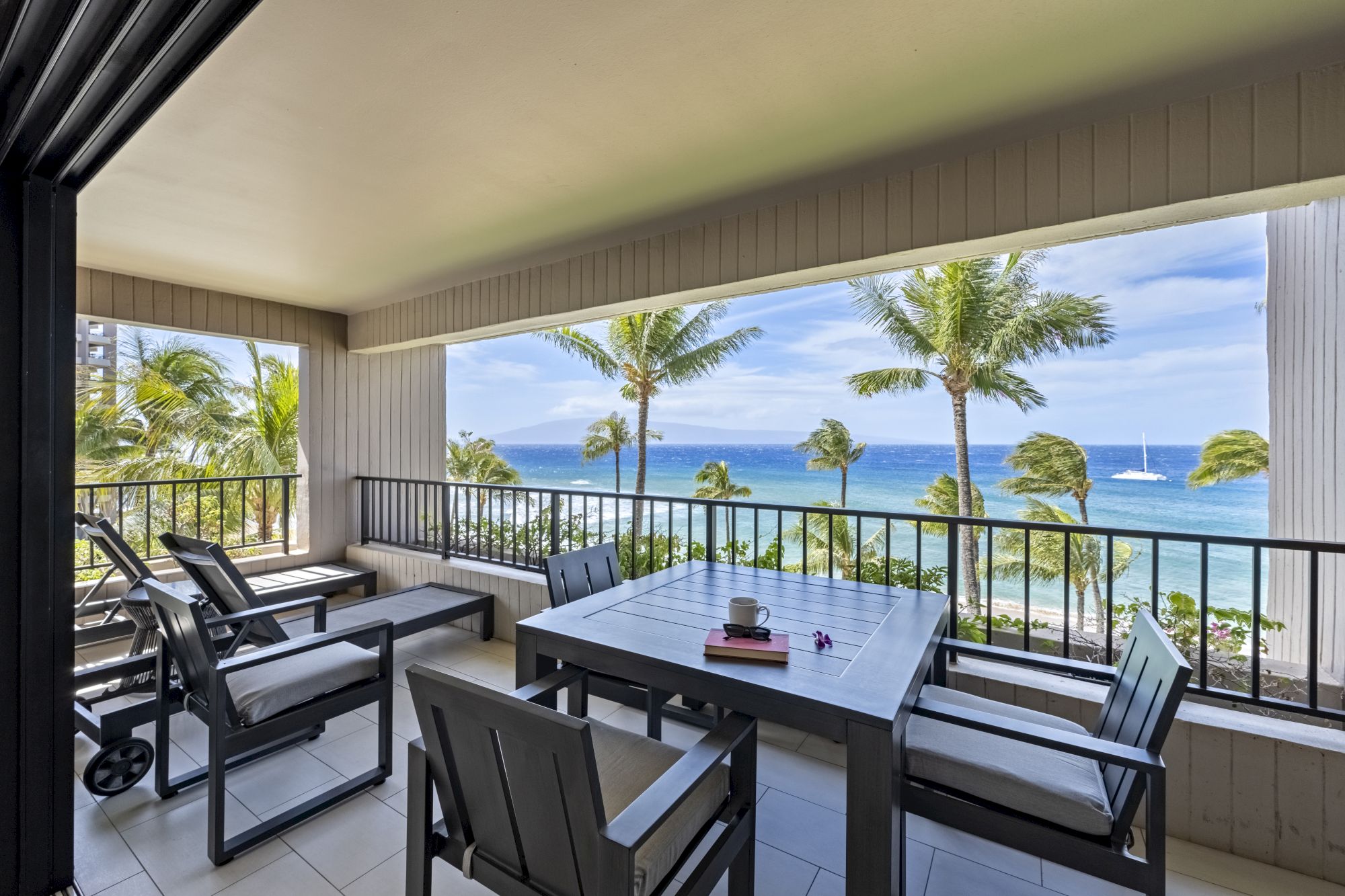 A covered patio with outdoor furniture overlooks a scenic tropical beach with palm trees, clear blue skies, and a boat on the horizon.