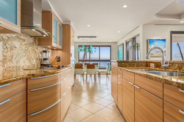 This image shows a modern kitchen with wooden cabinetry, granite countertops, and a dining area with a table and chairs in the background.