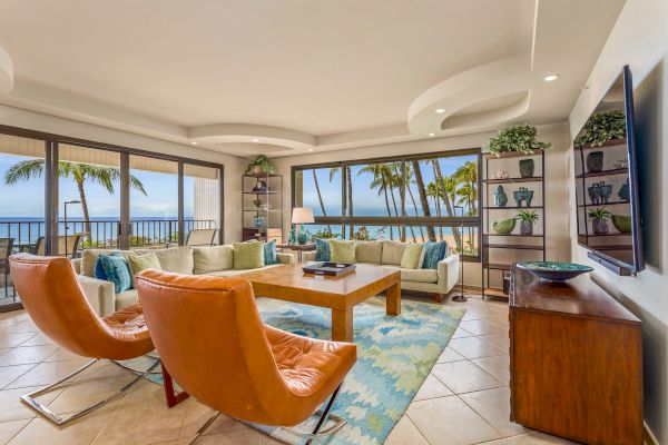 A living room with large windows showing an ocean view, modern furniture including orange chairs, a wooden table, and decorative shelves.