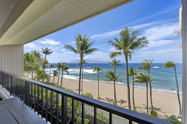 A beachfront balcony view featuring a sandy beach, palm trees, and a blue ocean with a sailboat in the distance, all under a clear, sunny sky.