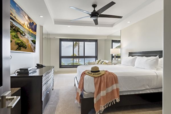 A modern bedroom with a large bed, ceiling fan, wall-mounted TV, dresser, and window view. A hat and blanket are placed on the bed.