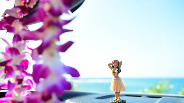 A hula girl figurine is on a car dashboard with a purple flower lei hanging nearby, against a backdrop of blue sky and ocean.