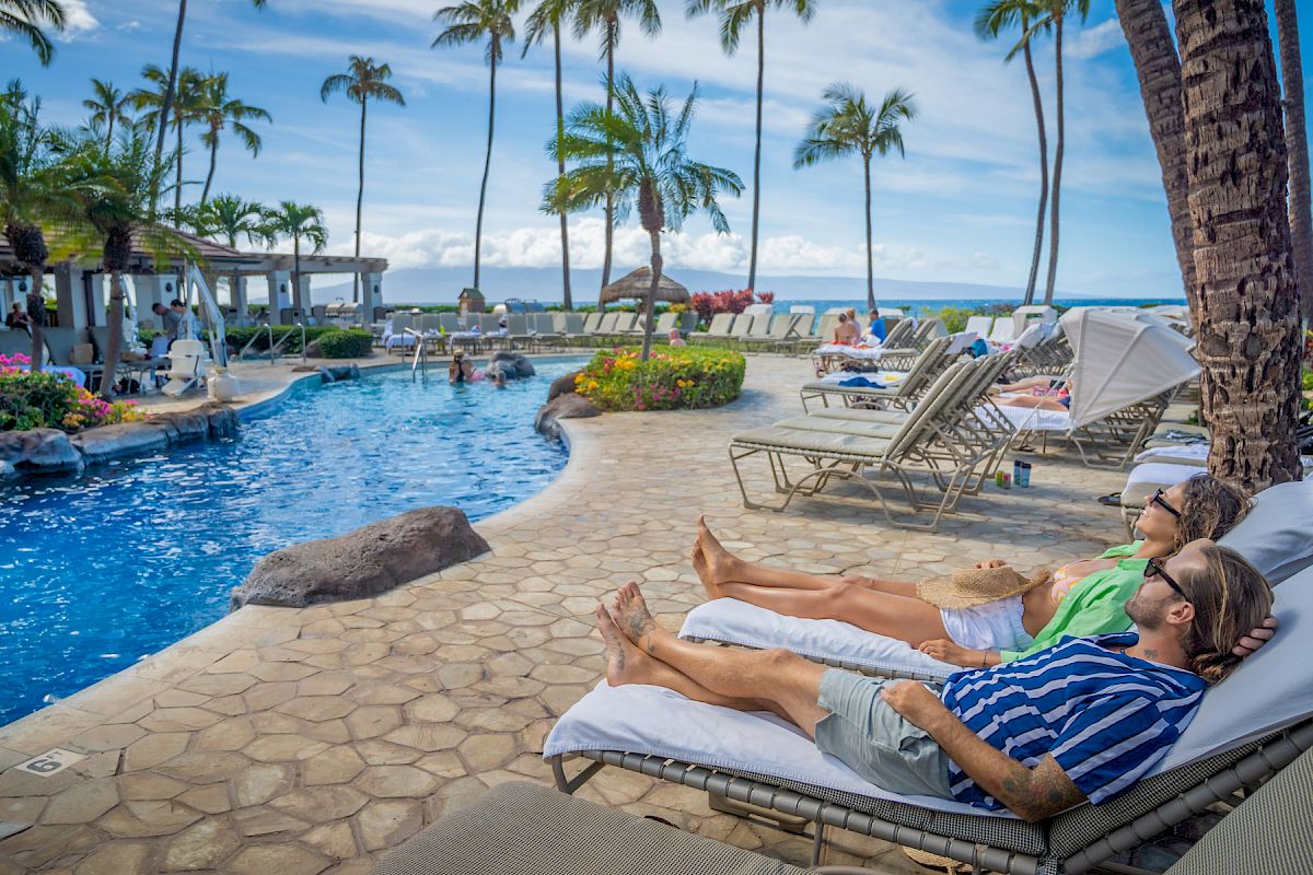 Two people are lounging on chairs by a pool, under palm trees with an ocean view and clear blue sky in the background.