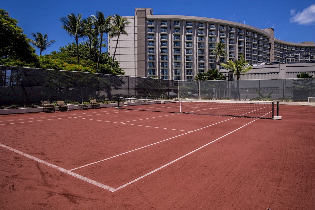 The image shows a clay tennis court with a hotel building in the background, surrounded by palm trees and clear blue skies, ending the sentence.