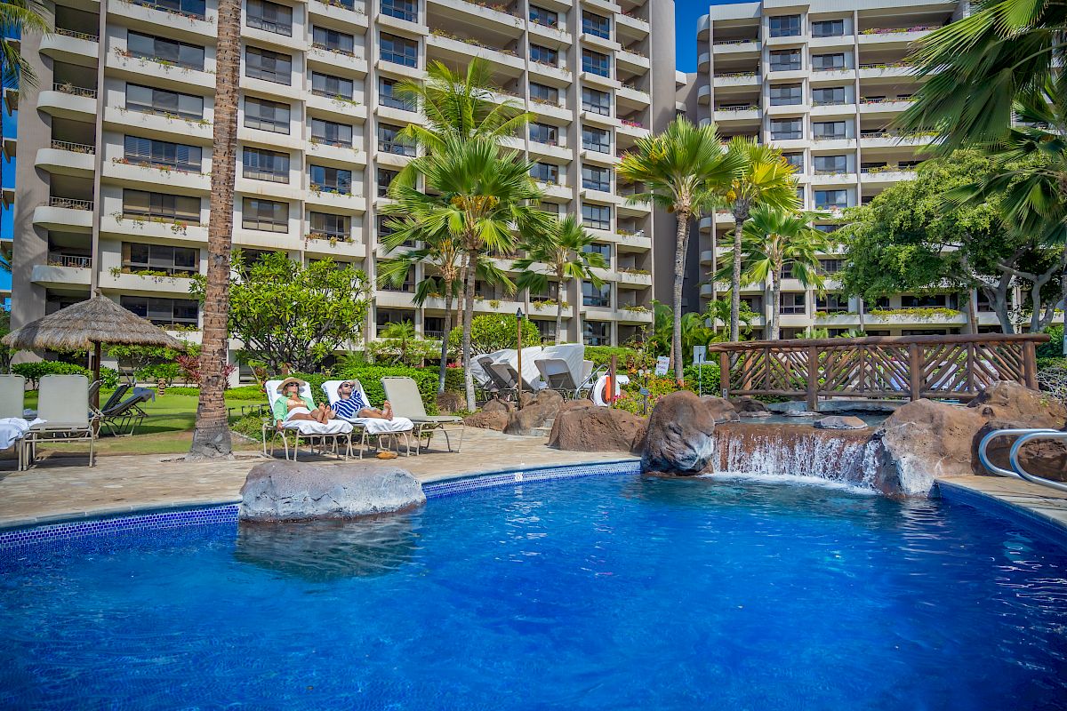 A serene pool area with lounge chairs, palm trees, and a waterfall feature, set against the backdrop of a multi-story residential building.