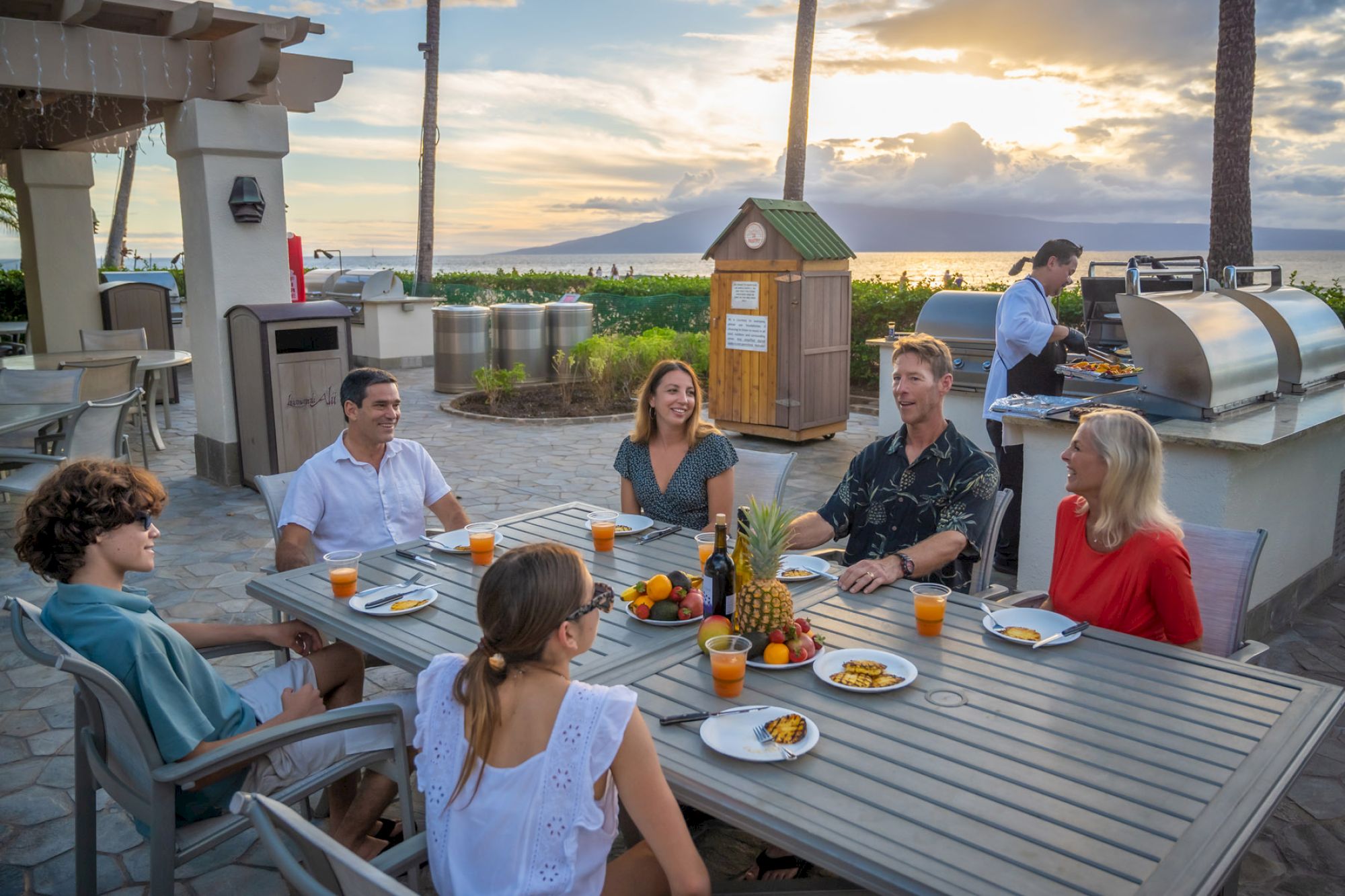 A group of people sit around a dining table outdoors, enjoying drinks and food, with a scenic, sunset beach view in the background.