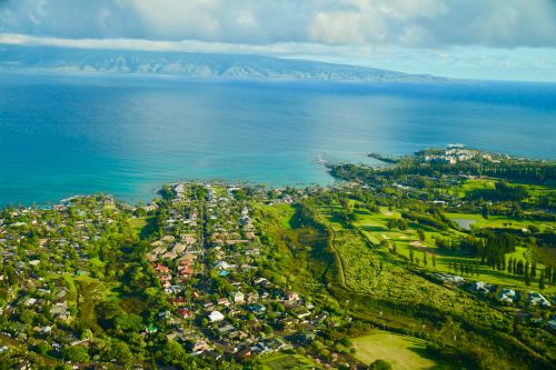 An aerial view of a coastal town with houses, lush greenery, and a golf course near the ocean, under a partly cloudy sky.