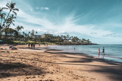 A scenic beach with people walking and playing in the sand and water, surrounded by palm trees and buildings in the background under a blue sky.