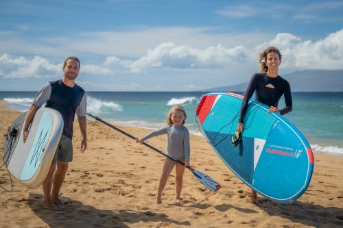 A family of three is standing on a beach, holding surfboards and paddles, with the ocean and sky in the background.